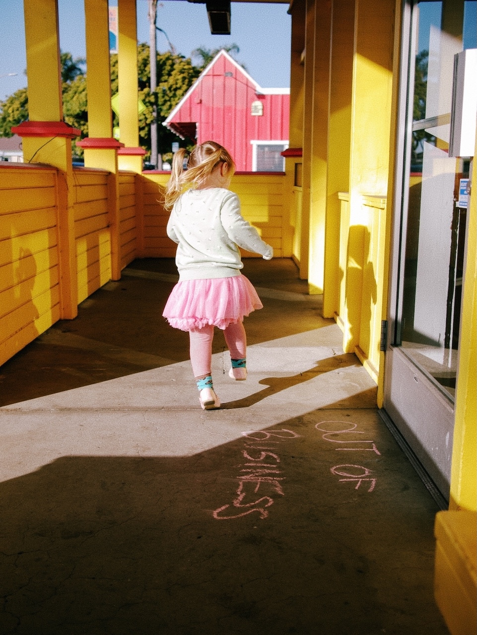 Brightly colored photo. A girl in a tutu skips past the door of a business. On the ground in chalk is written “out of business”.