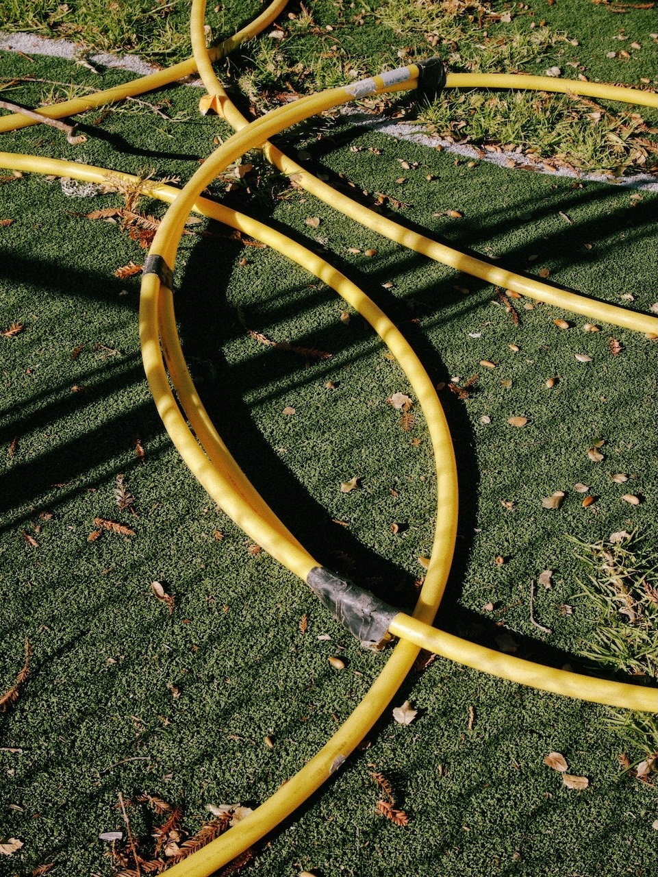 Color photograph of broken yellow hula hoops lying on fake grass. A shadow of a chain link fence covers them.